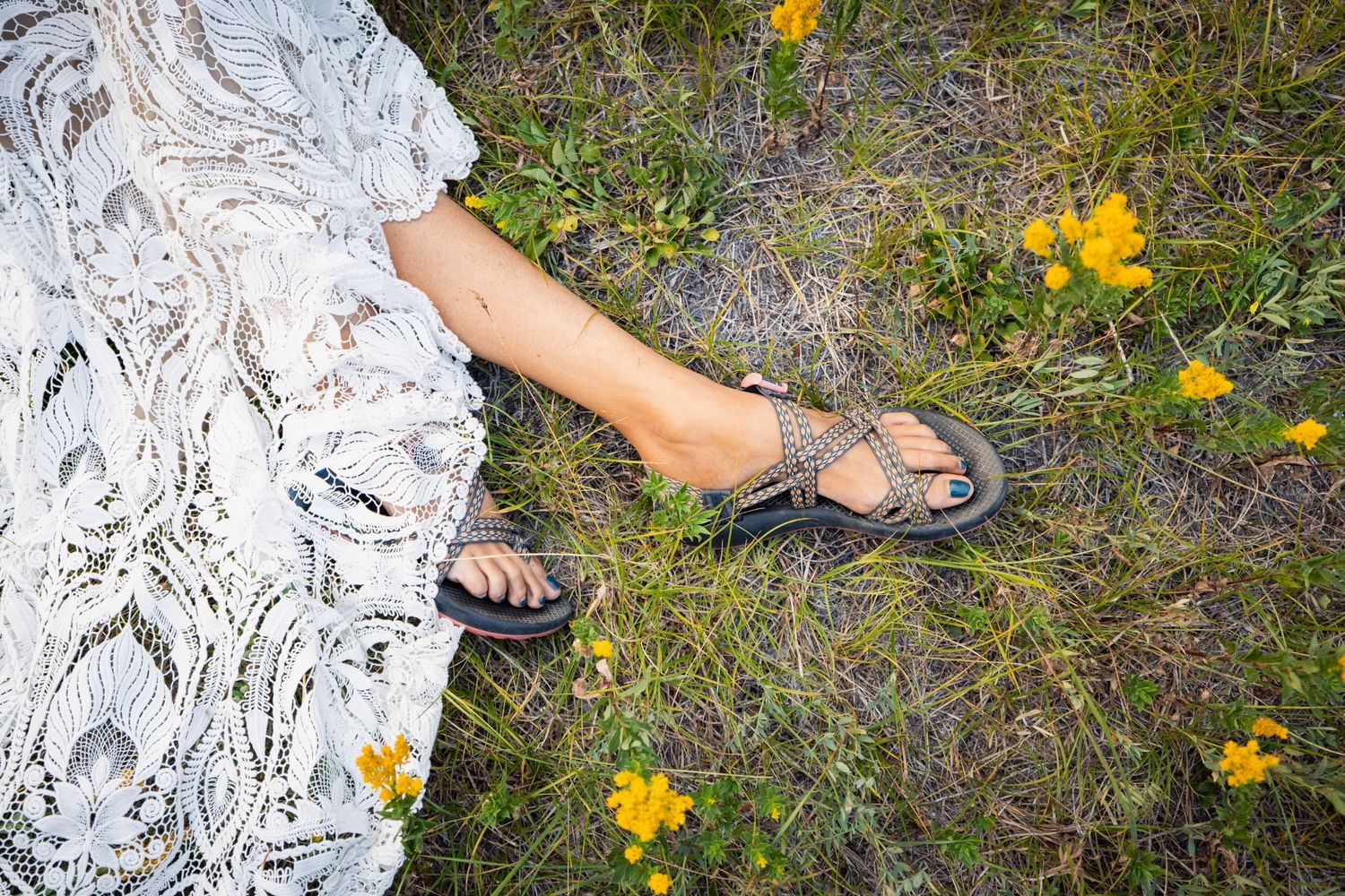 Feet wearing gray sandals on grassy ground with scattered yellow wildflowers, wearing a white lace dress.