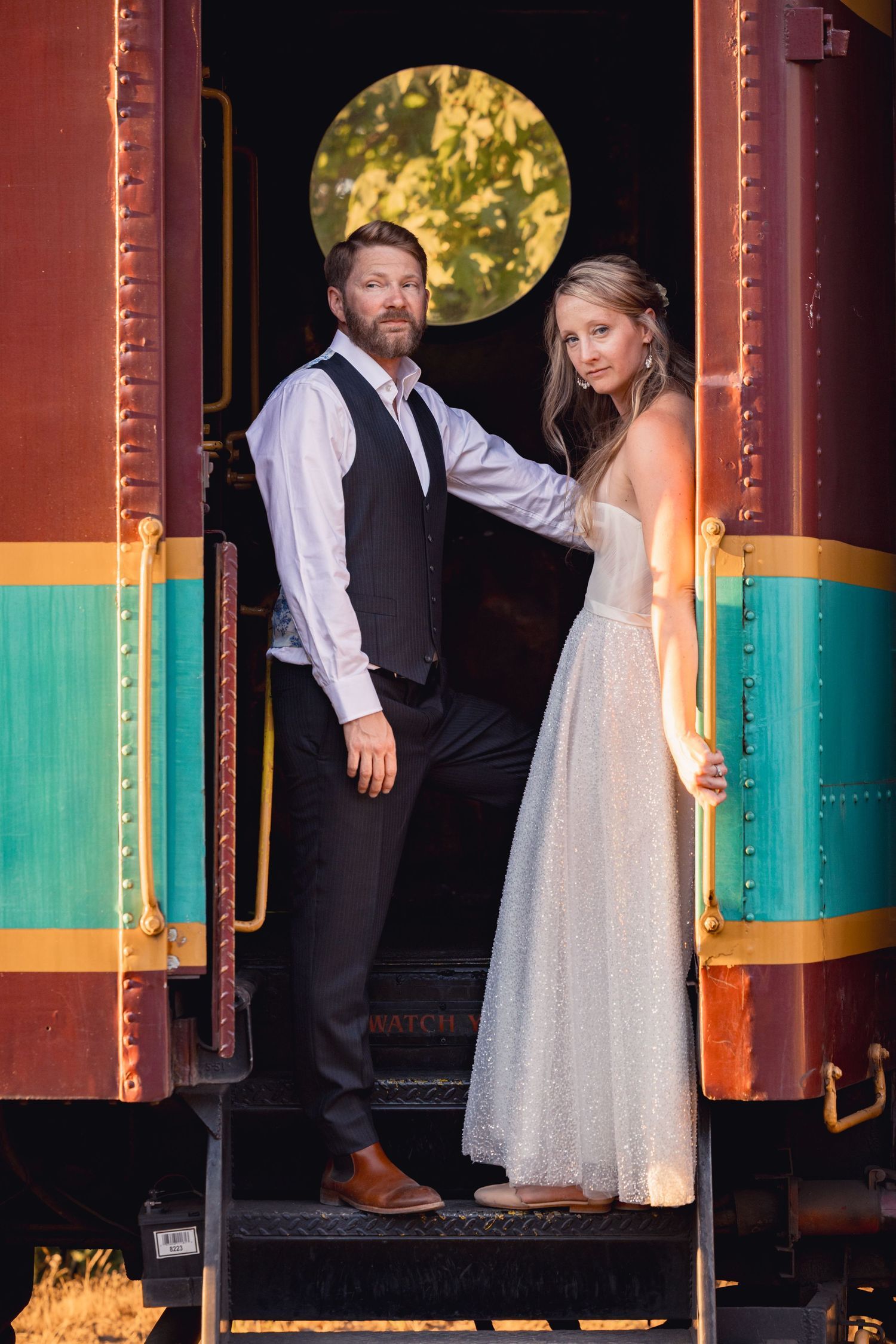 A couple poses on the steps of a vintage train car with colorful stripes, the bride wearing a sparkly white gown.