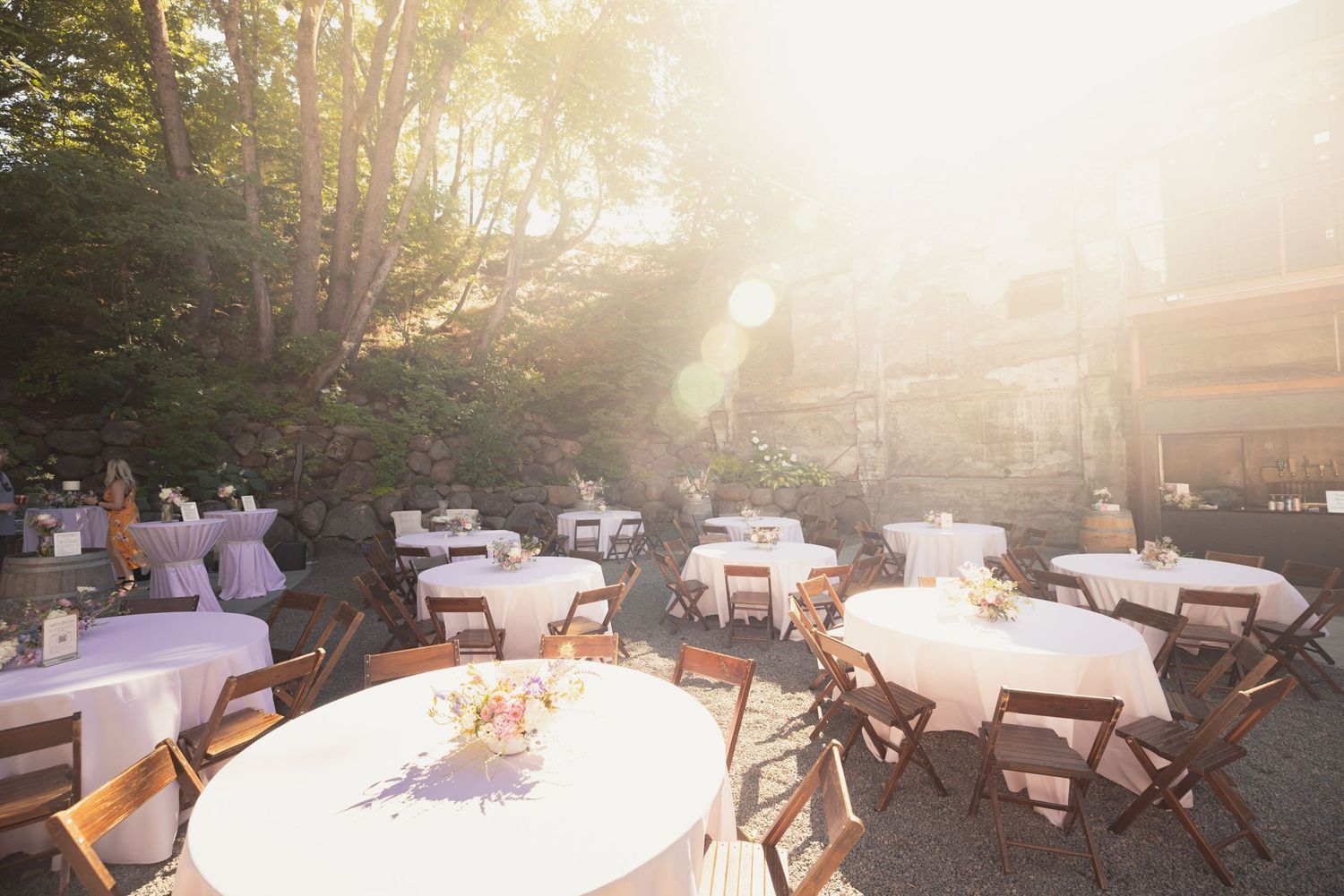 Outdoor wedding reception venue with white round tables and wooden chairs illuminated by warm sunlight through trees.