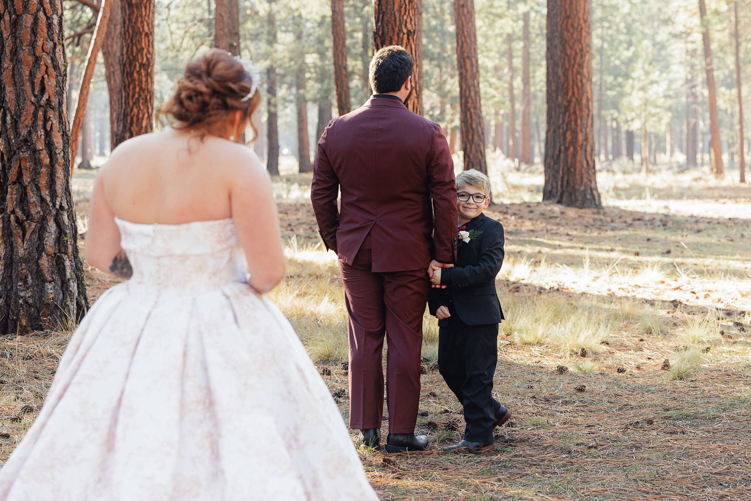 Couple in wedding attire stands among tall pine trees in a forest setting during an intimate ceremony.