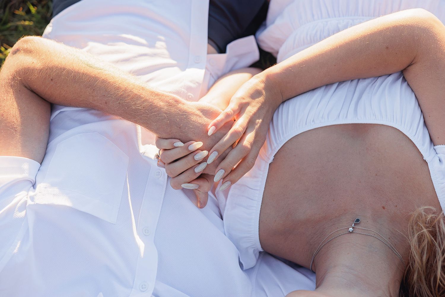Close-up of a couple holding hands while lying together, wearing white outfits in an outdoor setting.