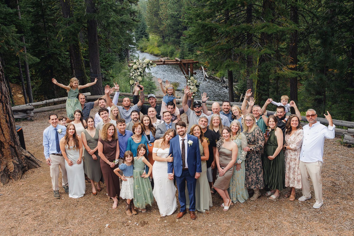Wedding party members pose together in a natural outdoor setting surrounded by tall evergreen trees.