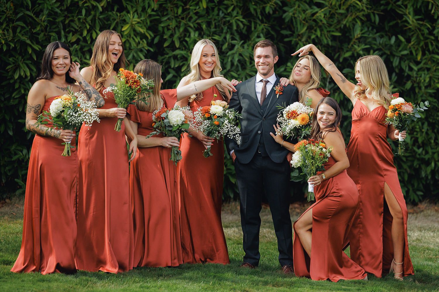 A wedding party poses outdoors with bridesmaids in flowing coral dresses holding colorful bouquets.