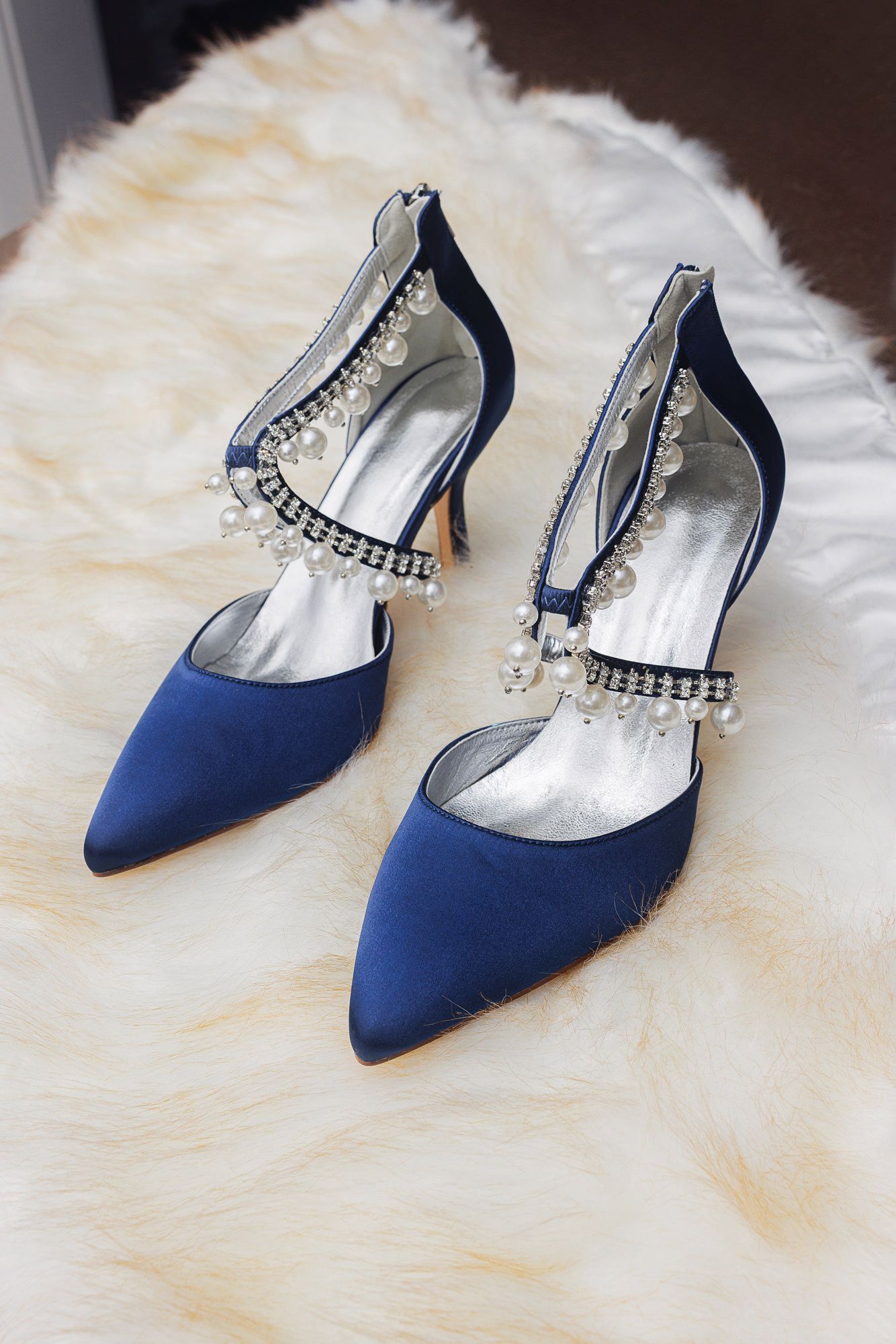 Navy blue satin kitten heel pumps with crystal embellished ankle straps displayed on white fur background.