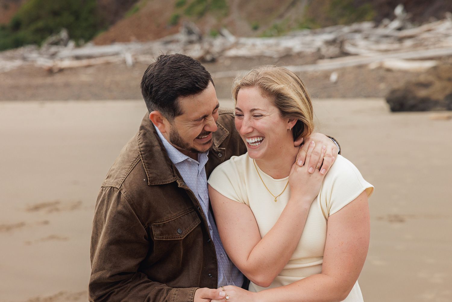 A happy couple laughs together on a sandy beach with driftwood, man in brown jacket, woman in white top.
