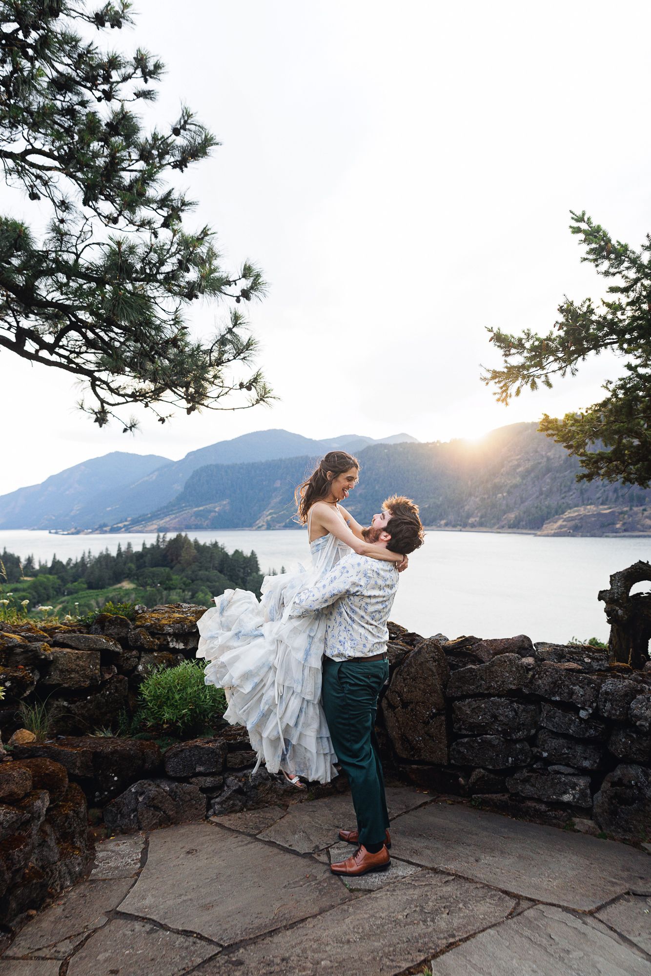 Groom lifts bride in white dress at scenic overlook with mountain lake and sunset backdrop.