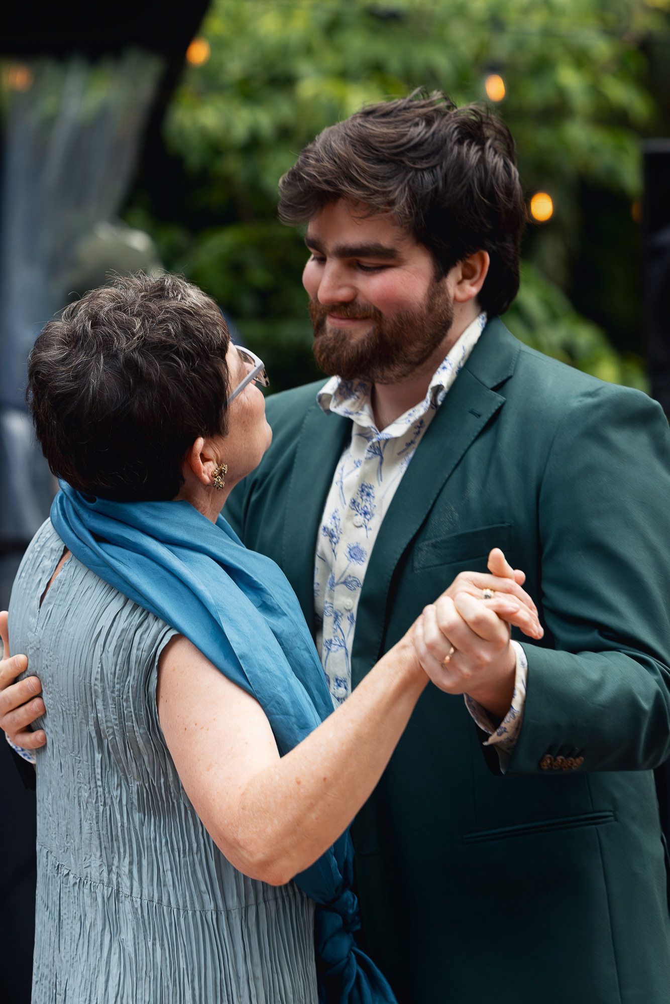 A young bearded man in a teal blazer shares a dance with an older woman in blue at an outdoor event.