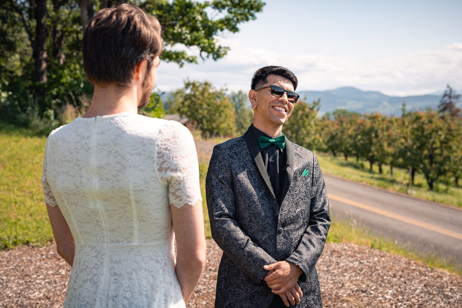 Wedding couple stands on a scenic mountain path surrounded by trees and distant blue mountains on a sunny day.