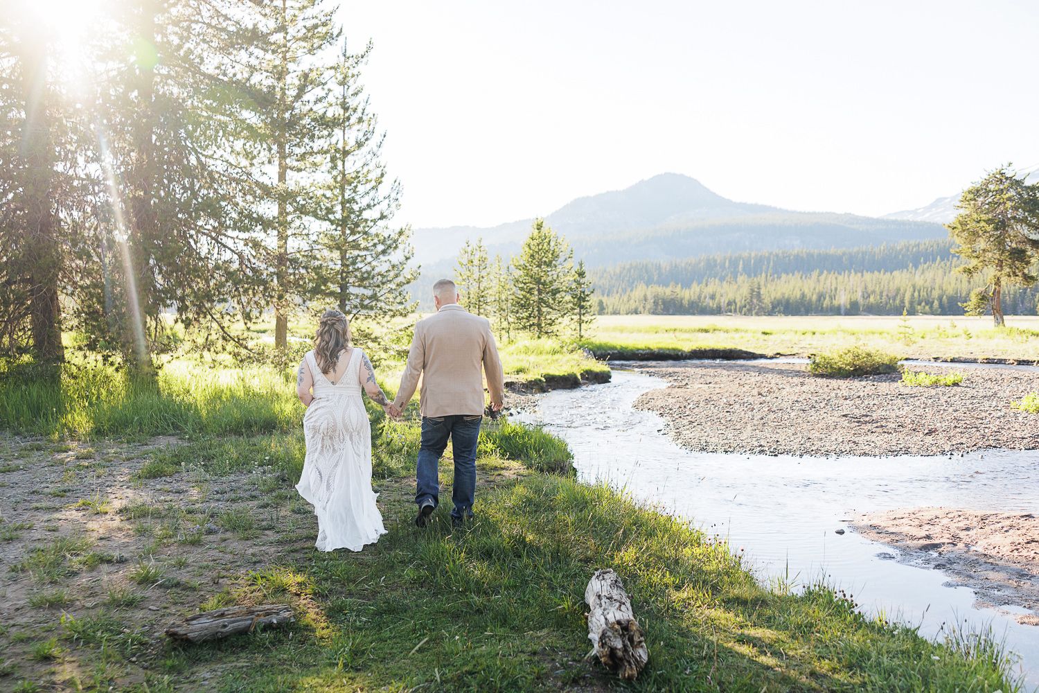 Two people stand near a scenic mountain river surrounded by evergreen trees in a natural landscape setting.