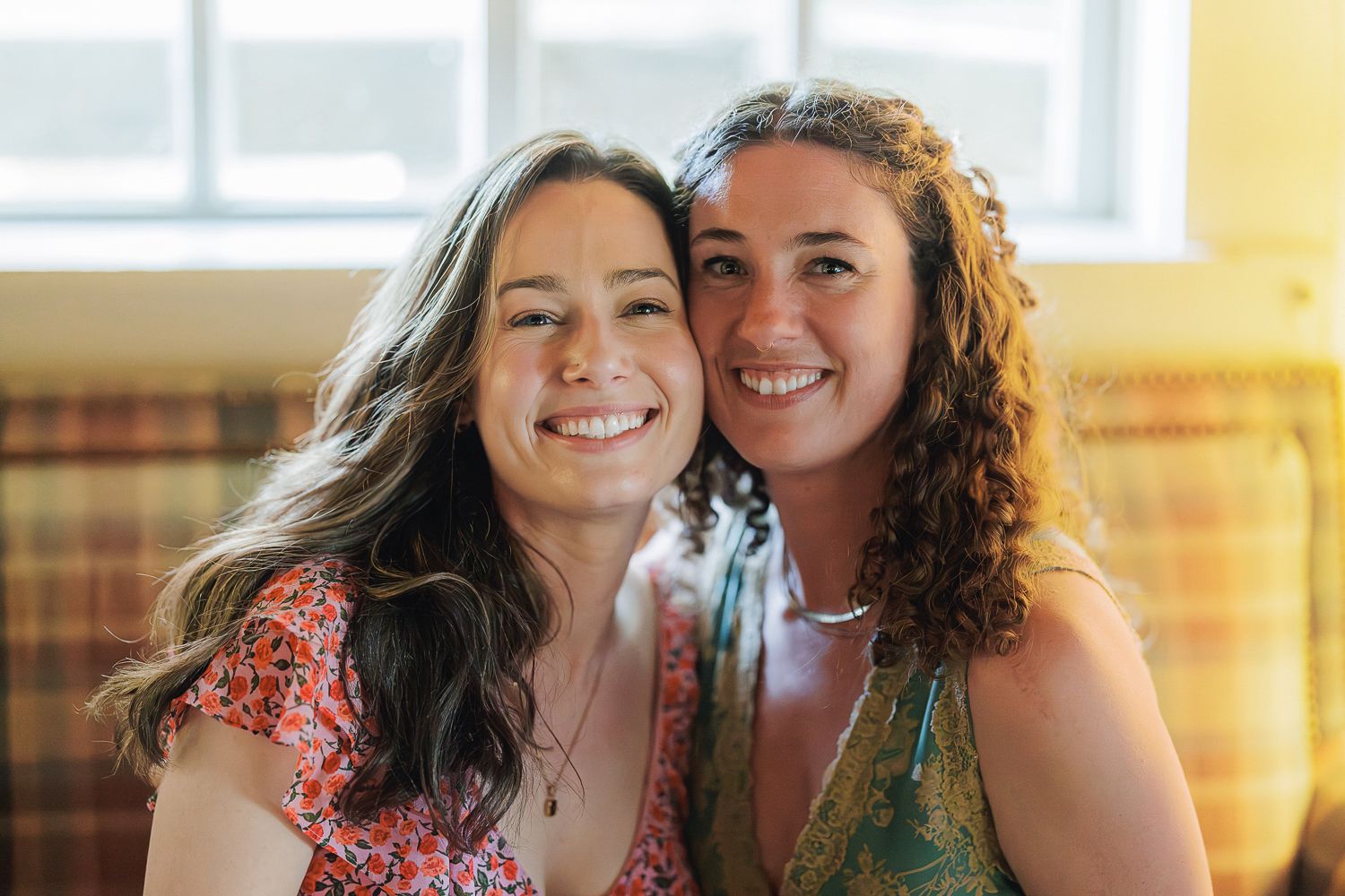 Two friends smile together in a warmly lit room wearing colorful floral dresses.