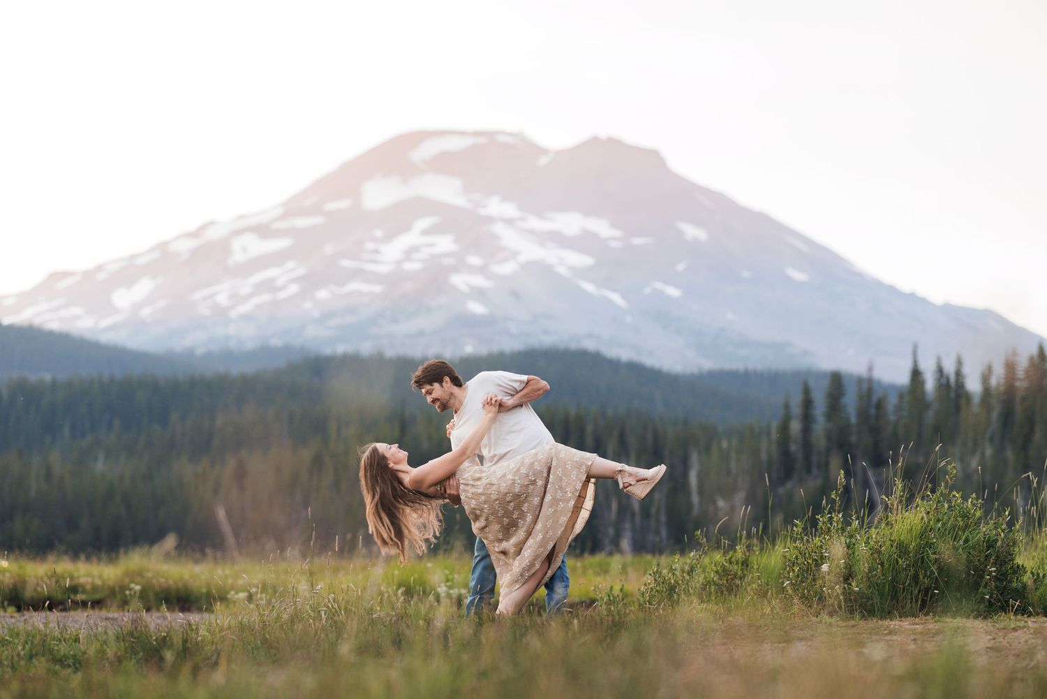 Couple dancing in a romantic outdoor setting with majestic snow-capped mountain and evergreen forest in the background.