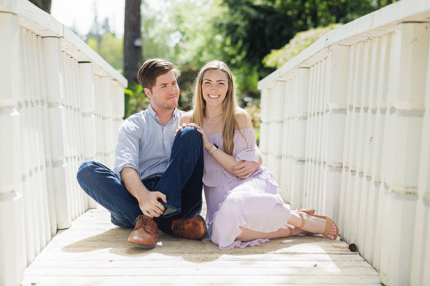 Couple sitting together on a white wooden bridge, smiling outdoors in a sunny park setting.