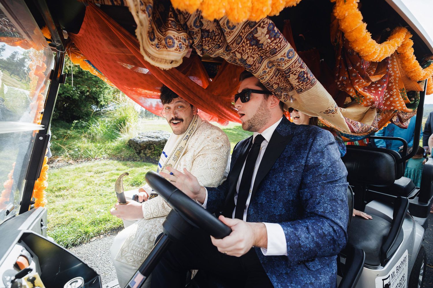 Two men in formal attire ride in a decorated vehicle with colorful orange garlands overhead.