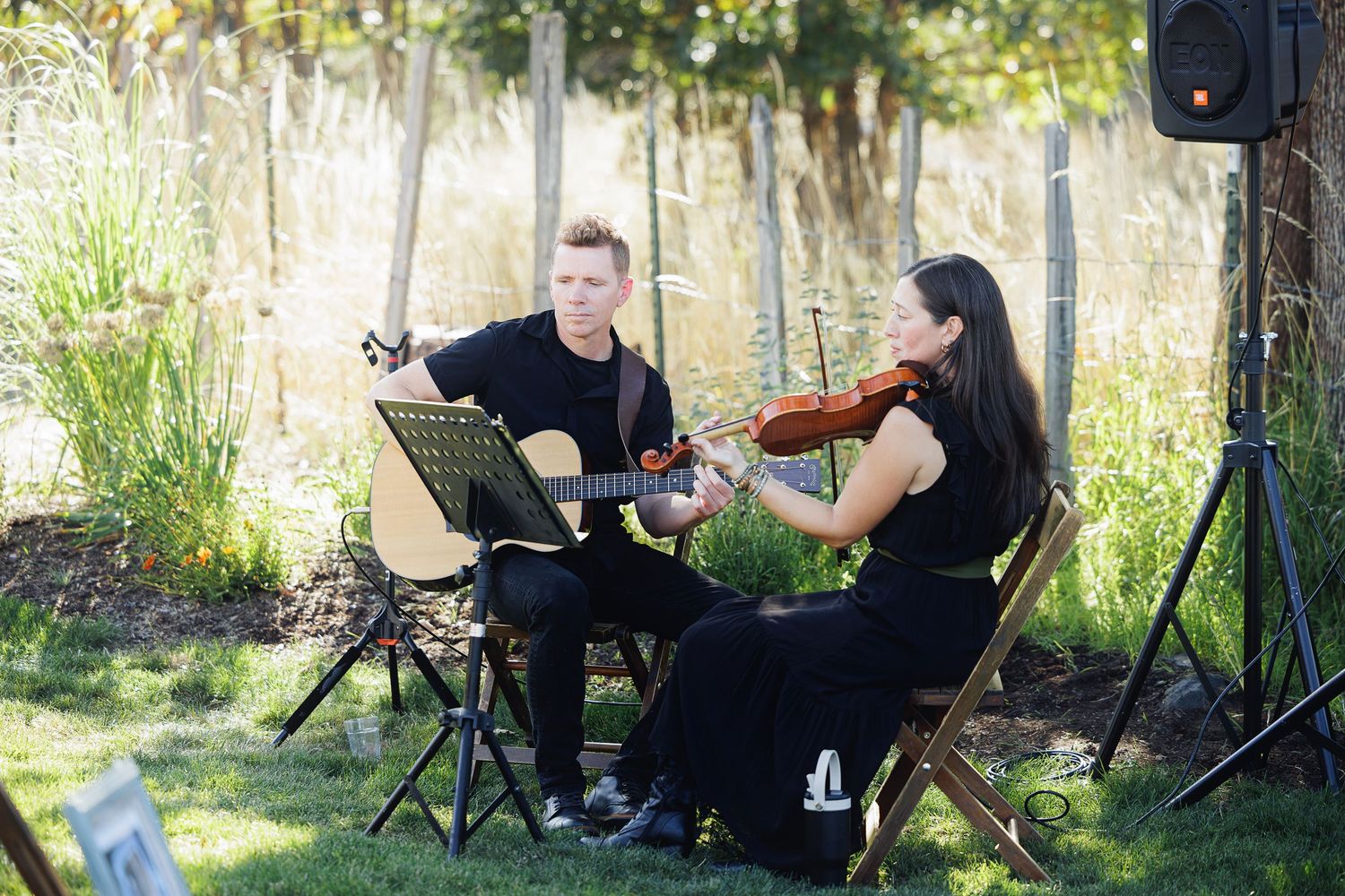 A guitarist and violinist duo dressed in black perform outdoors at a vineyard wedding ceremony.