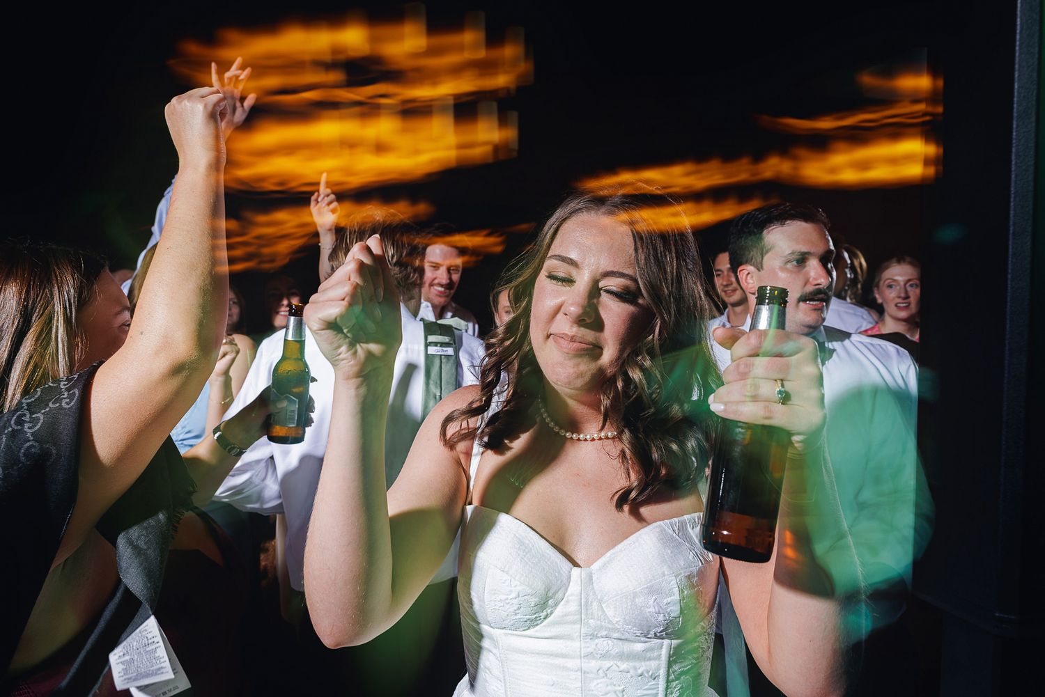 Bride in white strapless gown dancing joyfully with guests at wedding reception, holding beer bottle under warm lights.