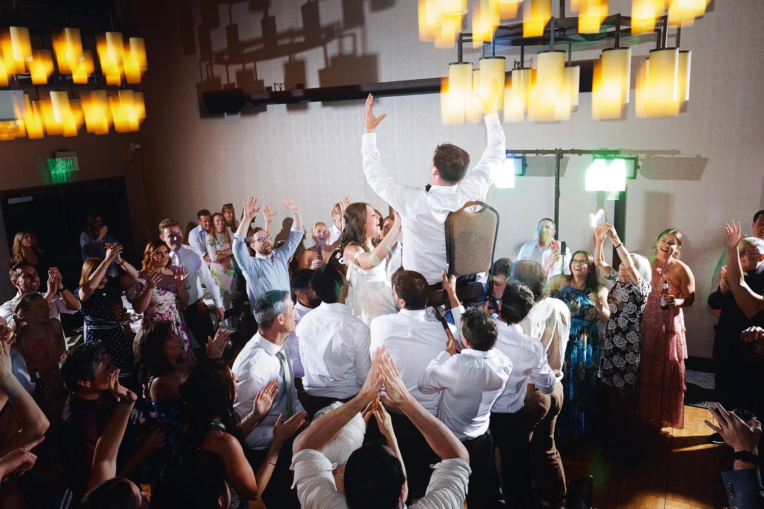 Wedding reception guests lifting bride and groom in celebration under warm chandelier lighting in elegant ballroom.