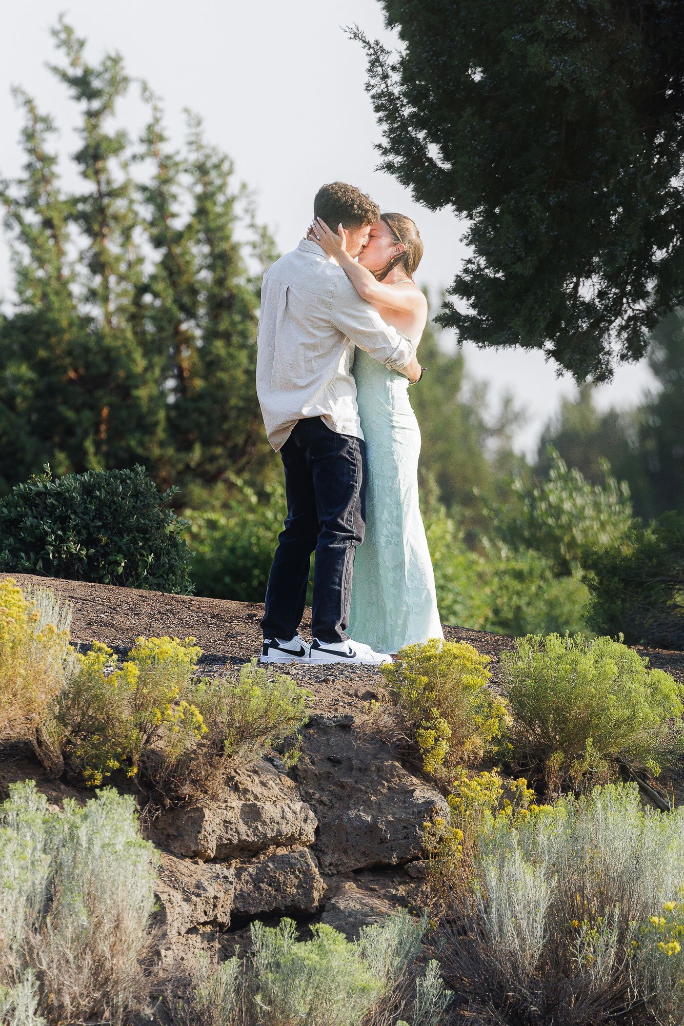 Couple embracing and kissing outdoors on rocky terrain surrounded by lush green trees and yellow wildflowers.