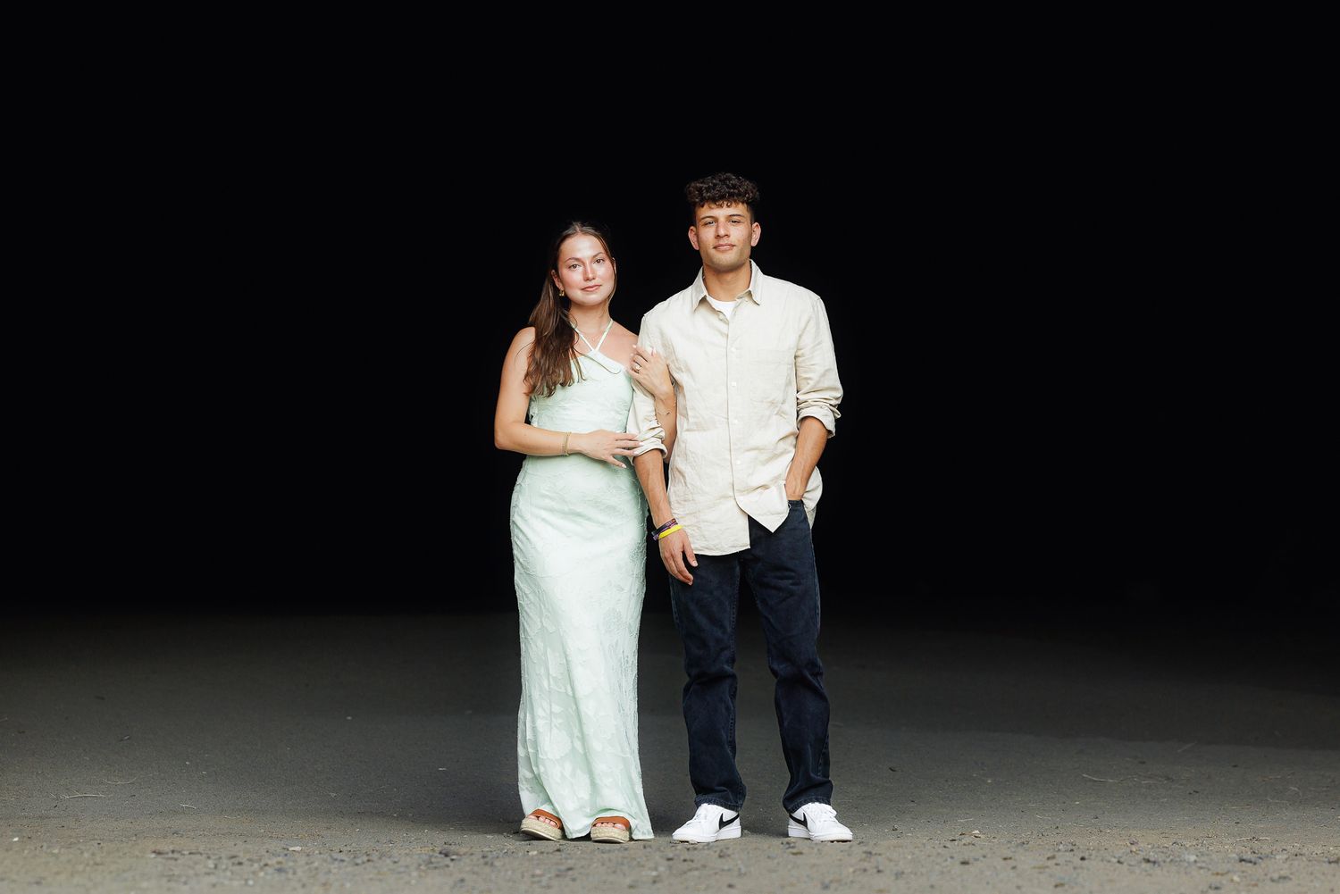 A young couple posing together against a dark background, woman in mint dress, man in beige shirt and dark jeans.