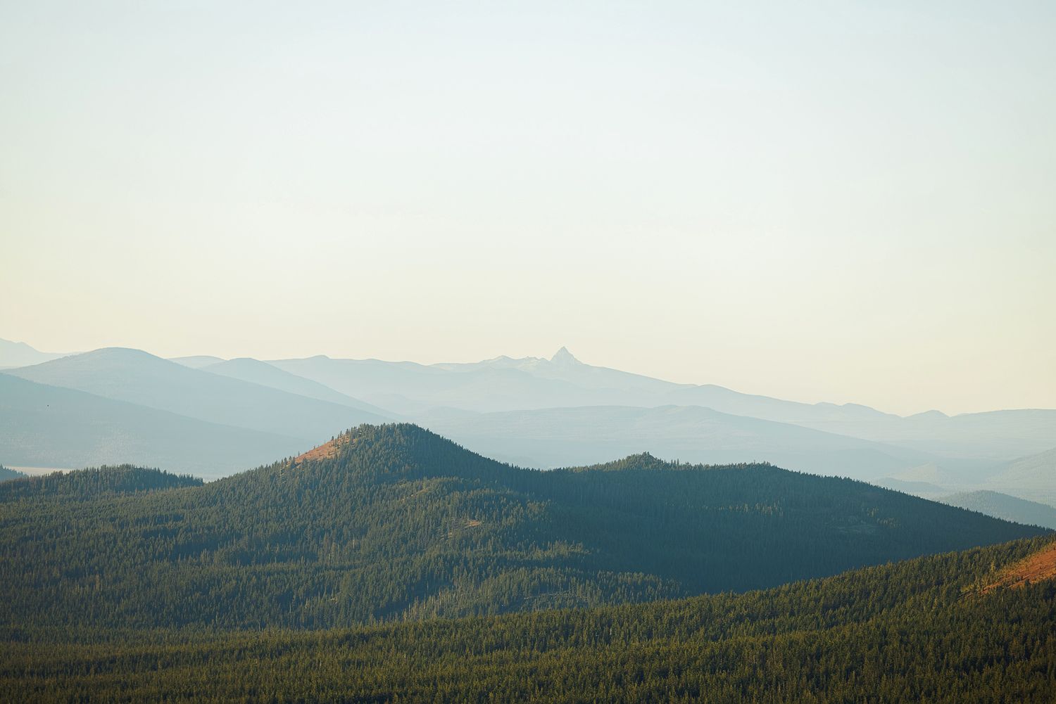 Layers of hazy blue mountain ridges fade into the distance behind a forested green hill under a soft, pale morning sky.