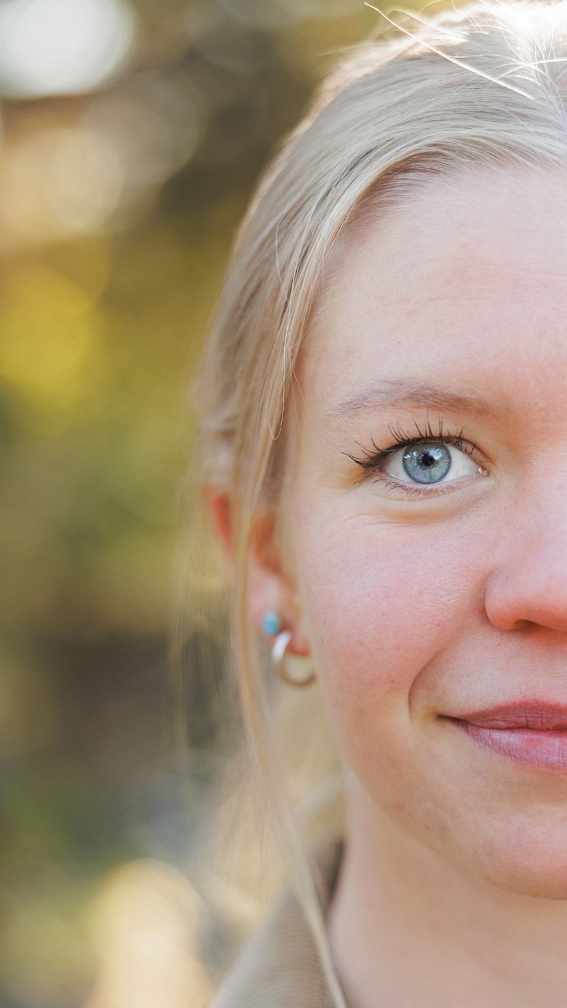 Close-up of half a smiling woman's face with blonde hair, blue eyes and a small colorful earring against a softly blurred autumn background.