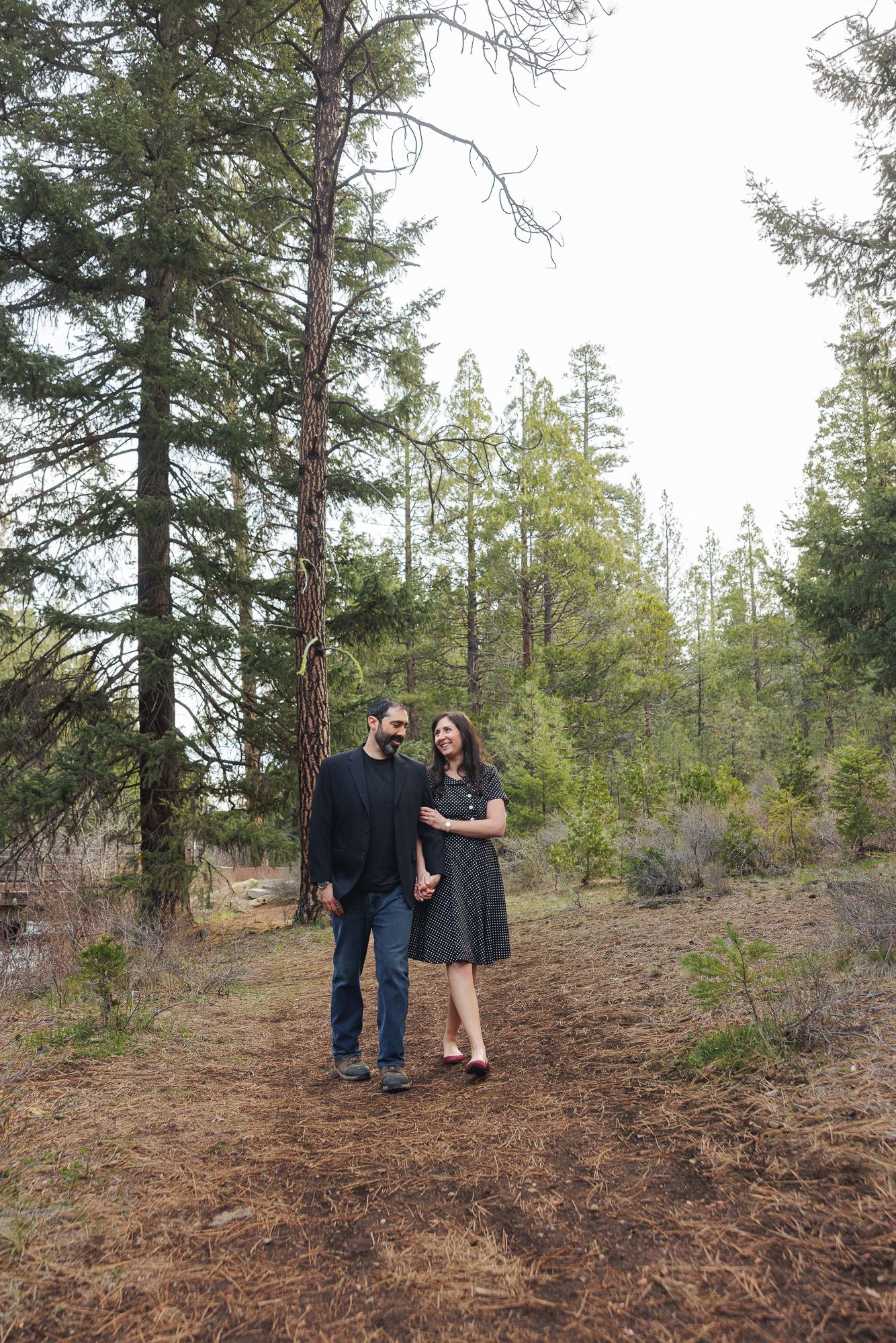 Couple holding hands and smiling at each other on a forest trail surrounded by tall pine trees.