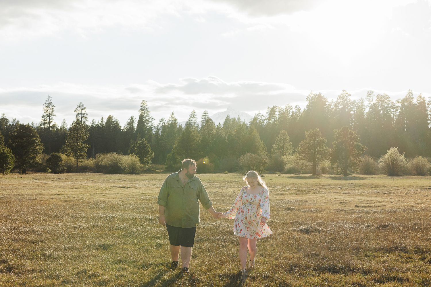 Couple holding hands walking through a sunlit meadow with trees in the background during golden hour.