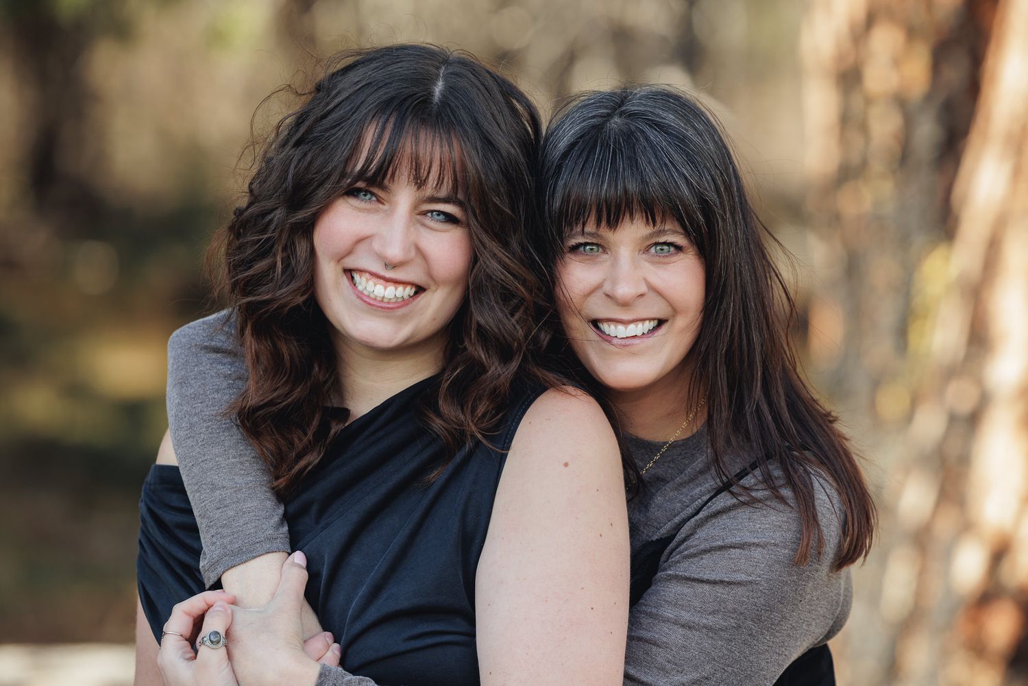 Two smiling brunette women with bangs hugging outdoors among trees in a warm, candid portrait photo.