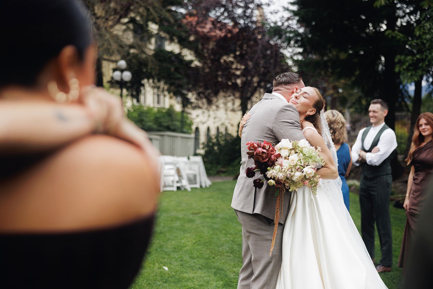 Bride and groom share first kiss at outdoor wedding ceremony on lush green lawn with guests watching.