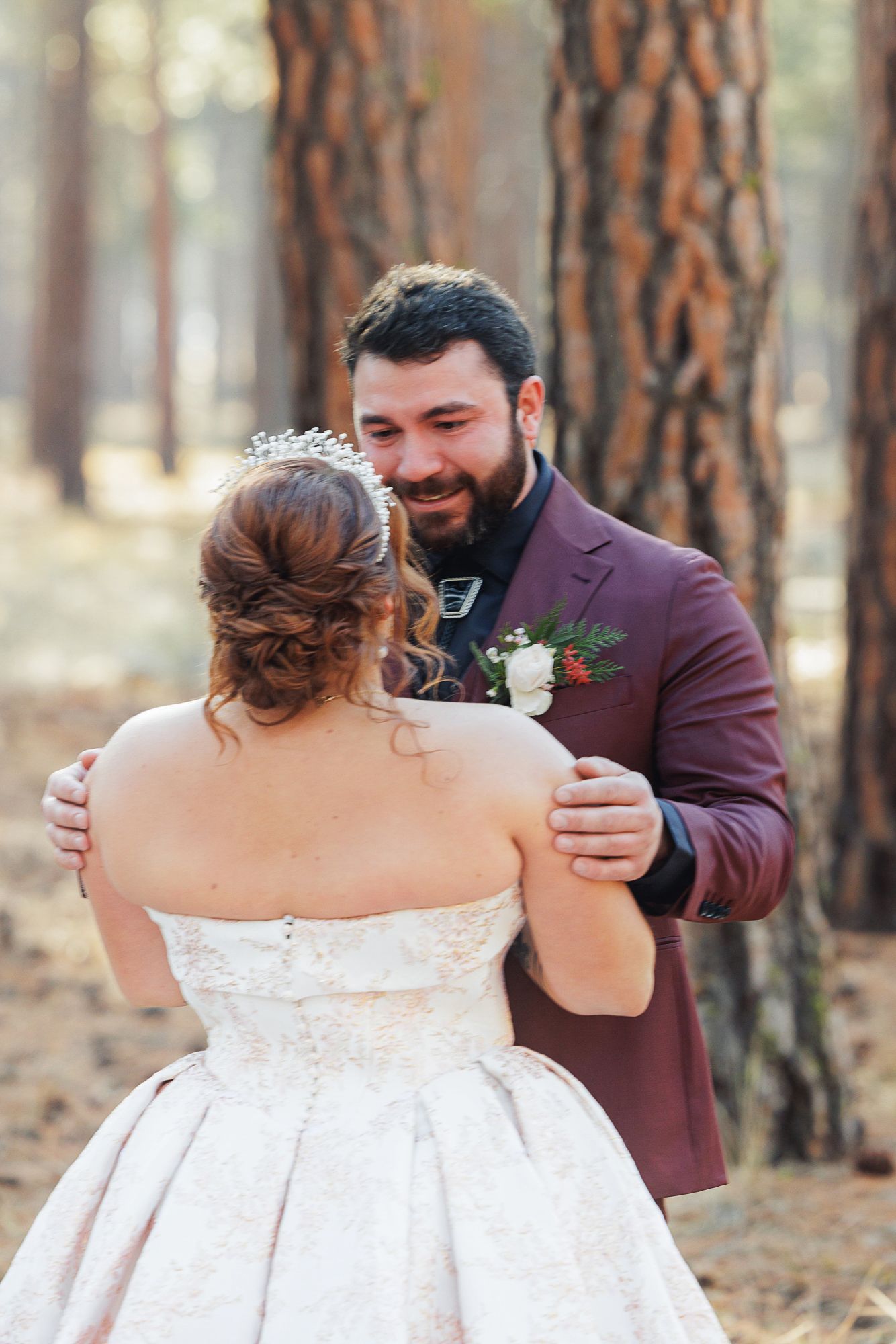 A romantic wedding moment captured in a pine forest with a burgundy suit jacket and white bridal gown.