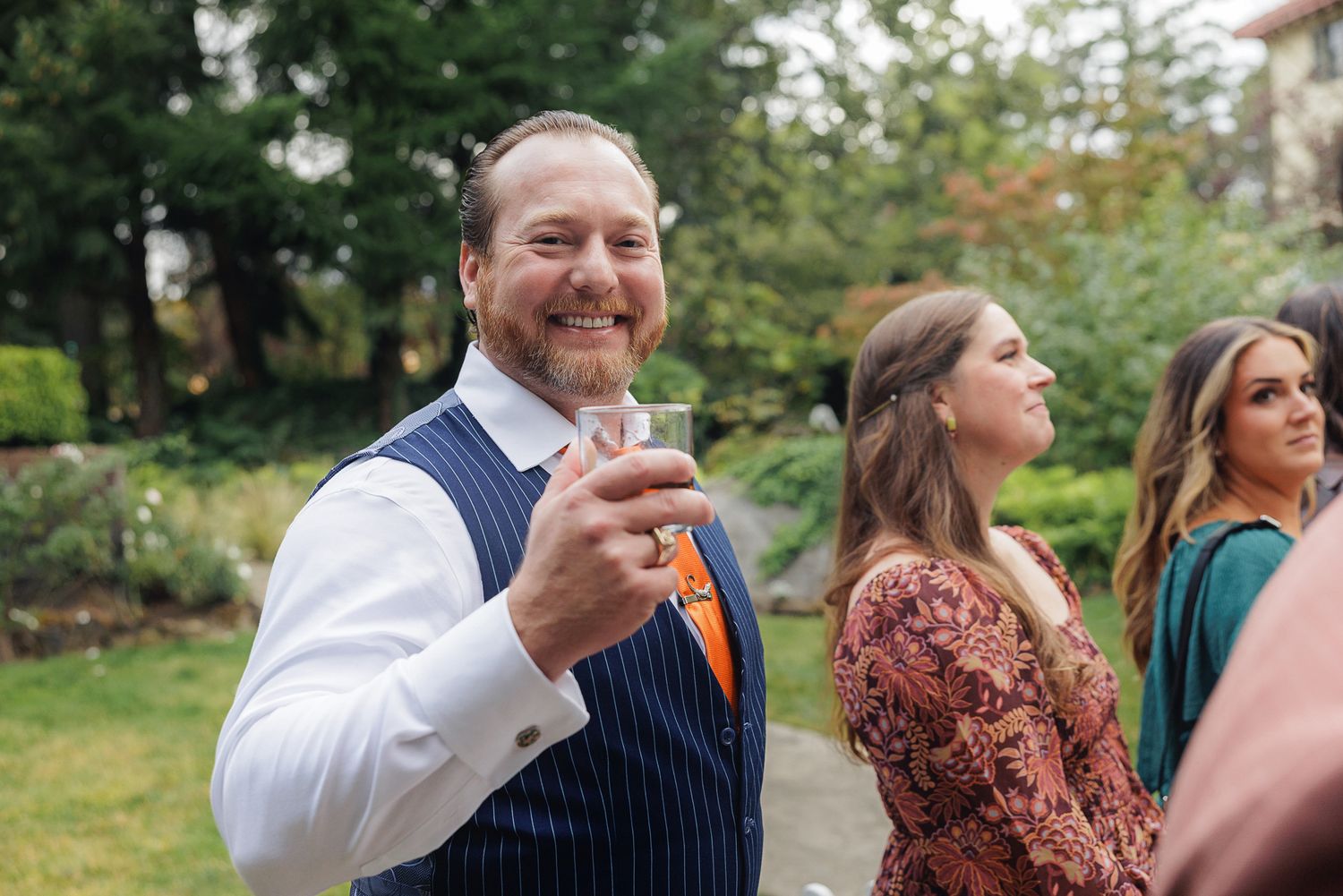 Wedding guests laugh and celebrate during an outdoor reception surrounded by greenery.