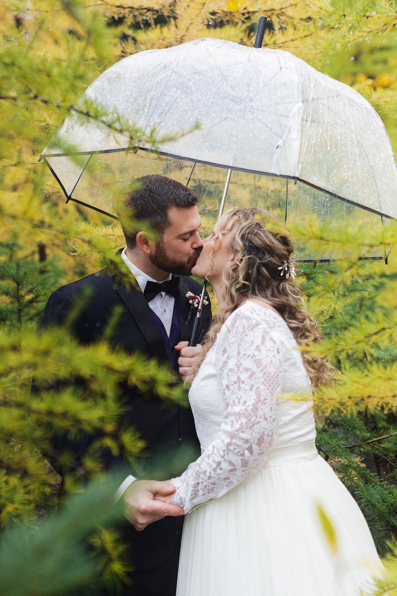 A romantic wedding couple shares intimate moments under a clear umbrella against a backdrop of autumn foliage.