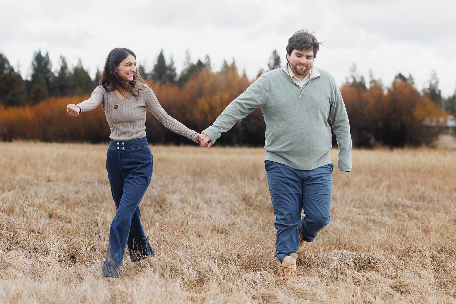 A couple playfully runs through a golden autumn field at sunset with evergreen trees in the background.