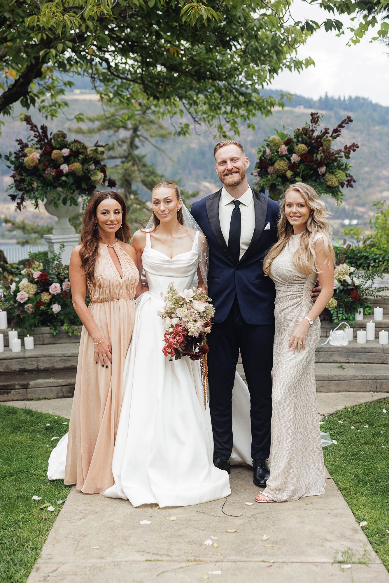 Bride and groom pose with two bridesmaids in blush gowns at outdoor wedding with floral arch backdrop.