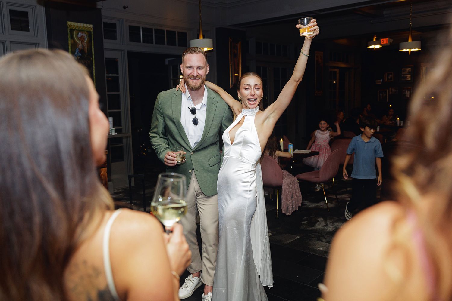 Bride in white gown raises glass with groom in green blazer at elegant wedding reception celebration.