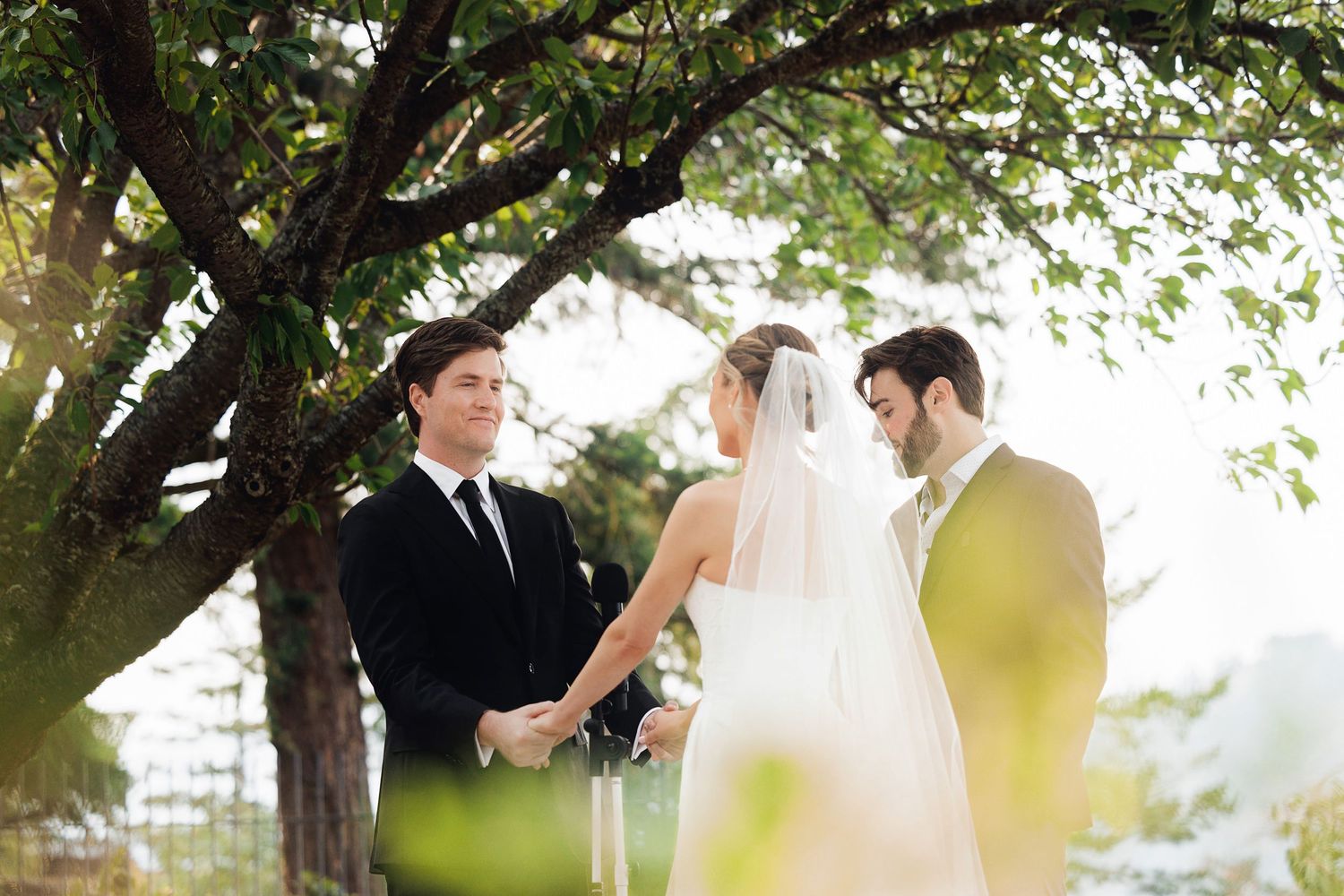 Couple exchanges vows outdoors under a tree, bride in white veil, groom in black suit, officiant present.
