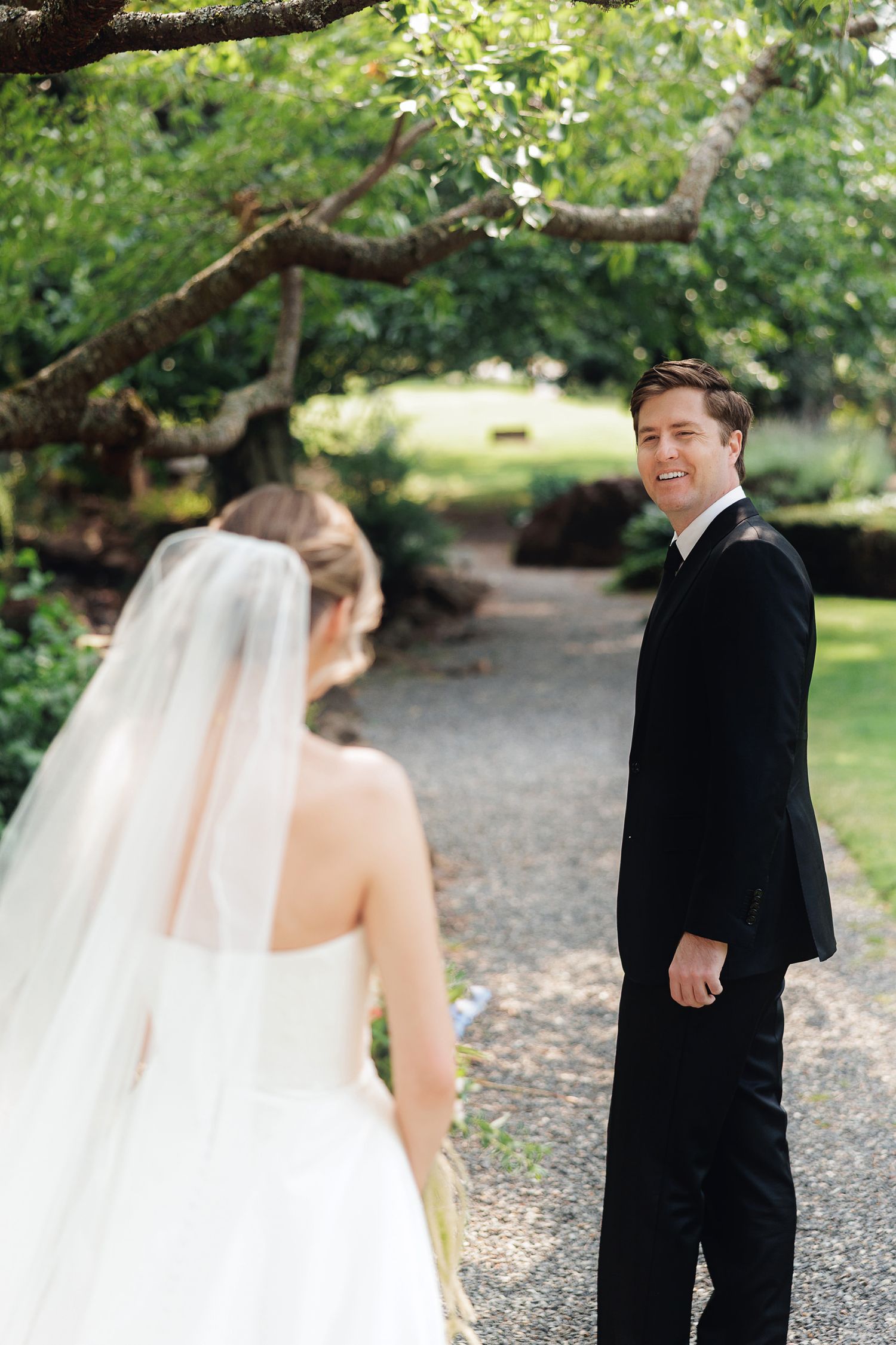 Groom in black tuxedo smiles at bride in white gown and veil on a tree-lined garden path.