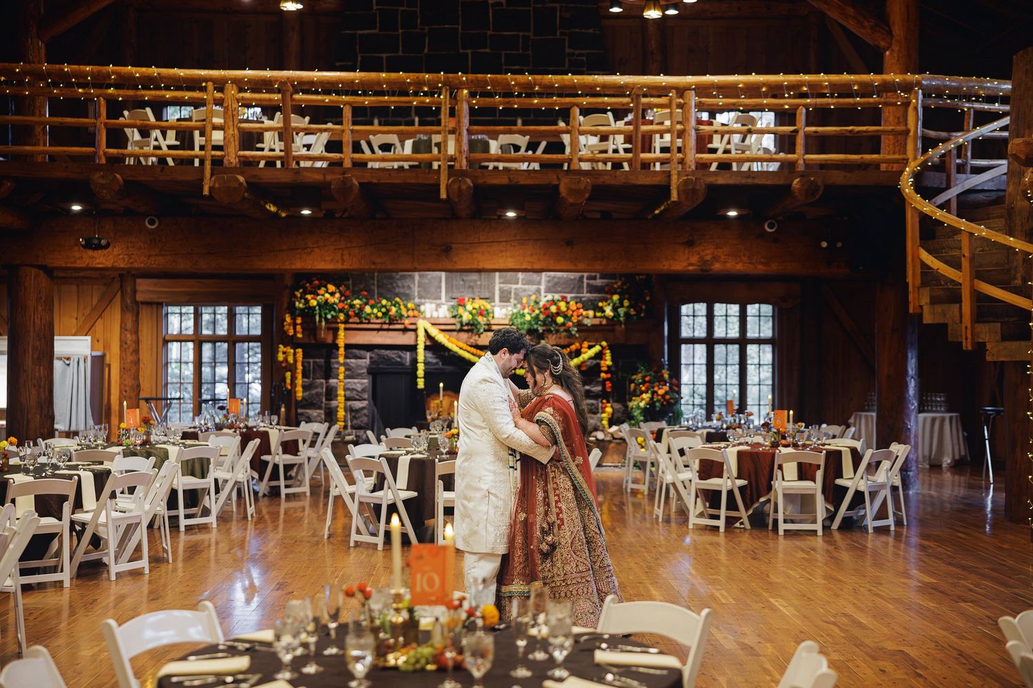 Indian couple shares first dance in rustic barn wedding venue with warm string lights and floral decorations.