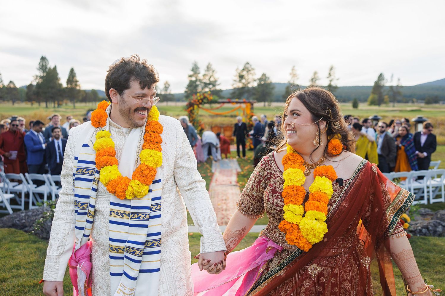 Couple in traditional Indian wedding attire with marigold garlands smiling joyfully after outdoor ceremony.