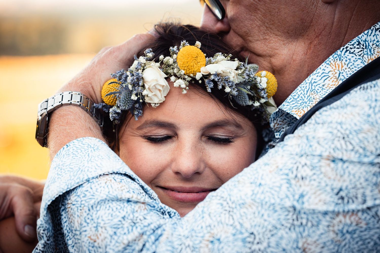 A person wearing a floral crown with yellow flowers and baby's breath embraces someone in soft, warm light.