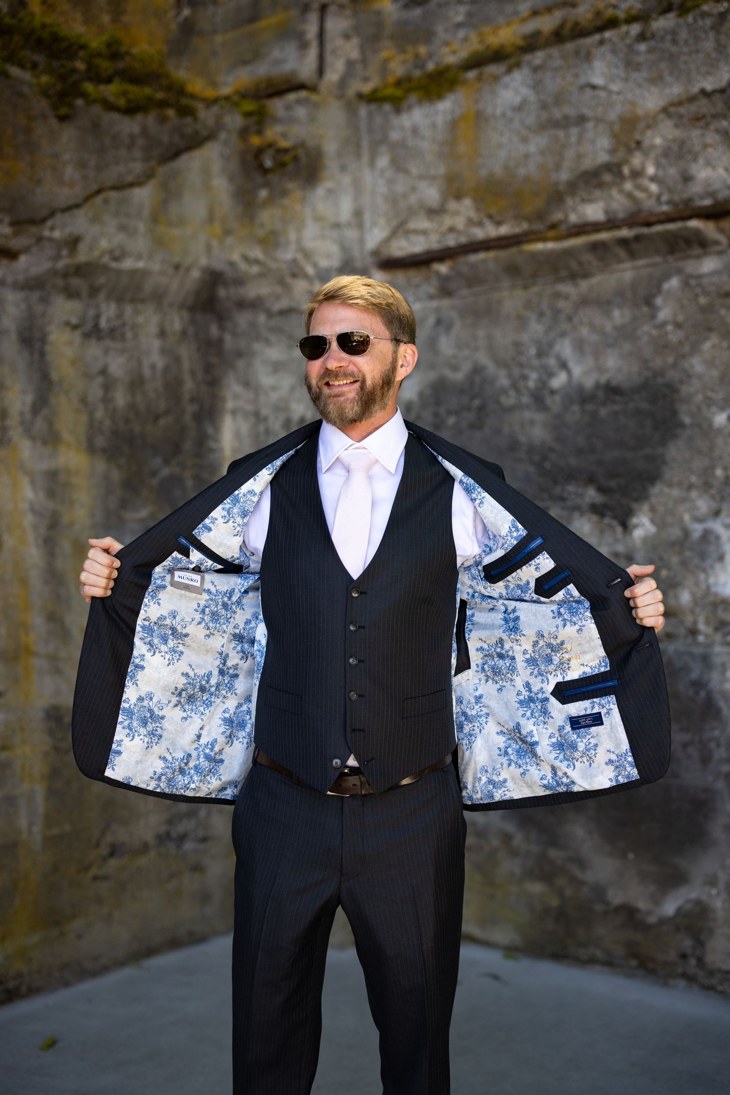 Person showing off stylish blue floral lining inside black suit jacket while wearing sunglasses against concrete wall.
