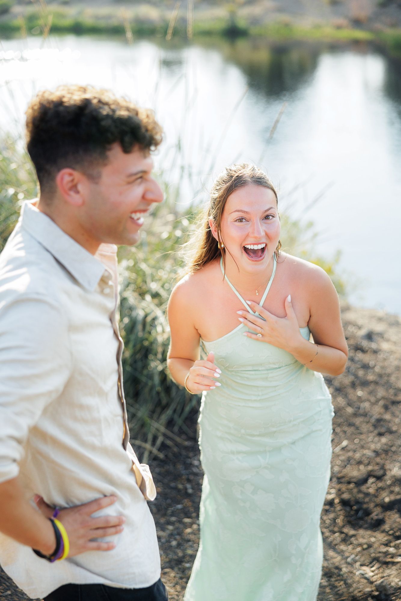 A joyful couple laughing together outdoors near a serene lake, woman in mint dress touching her chest with delight.