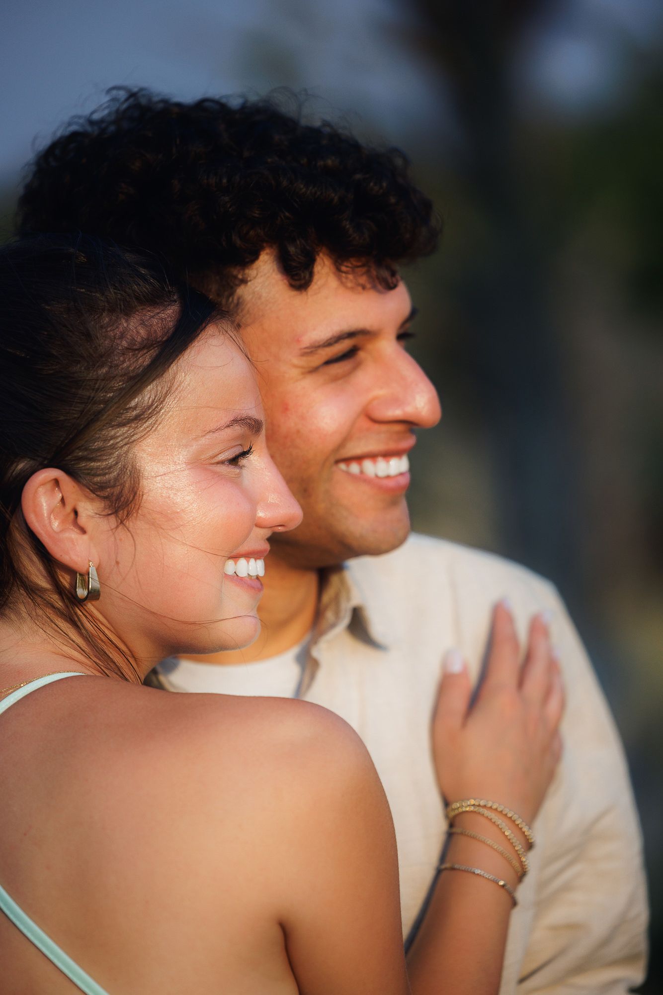 A smiling couple embraces outdoors in warm golden light, the woman wearing gold bracelets and a teal top.