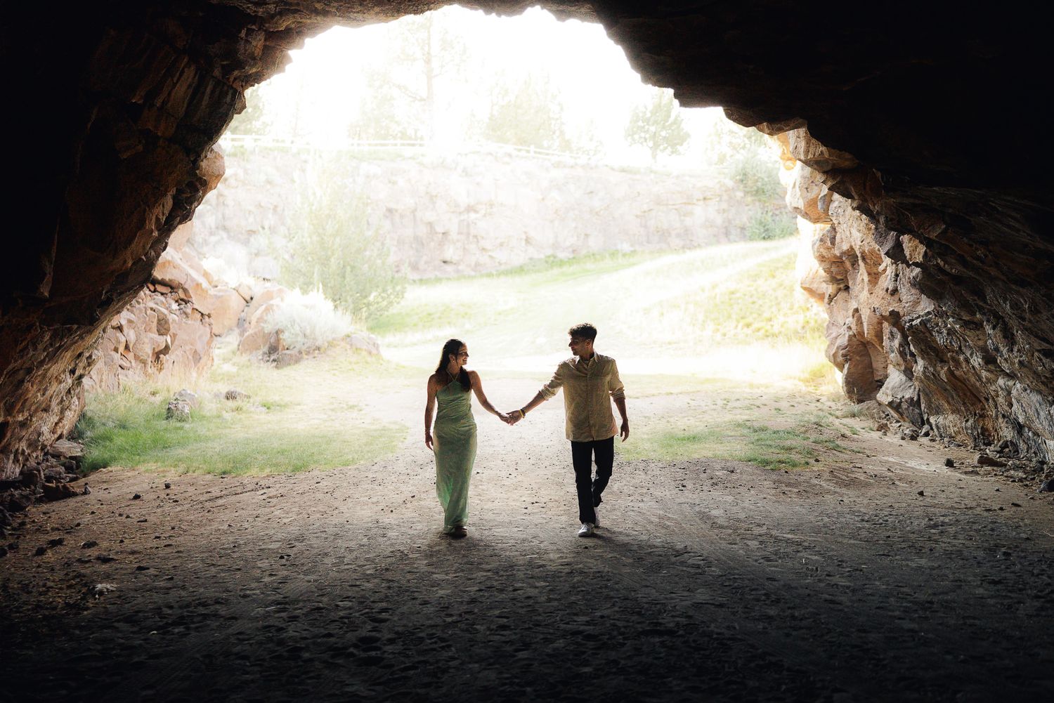 Couple holding hands silhouetted in cave entrance with sunlight and green landscape behind them.