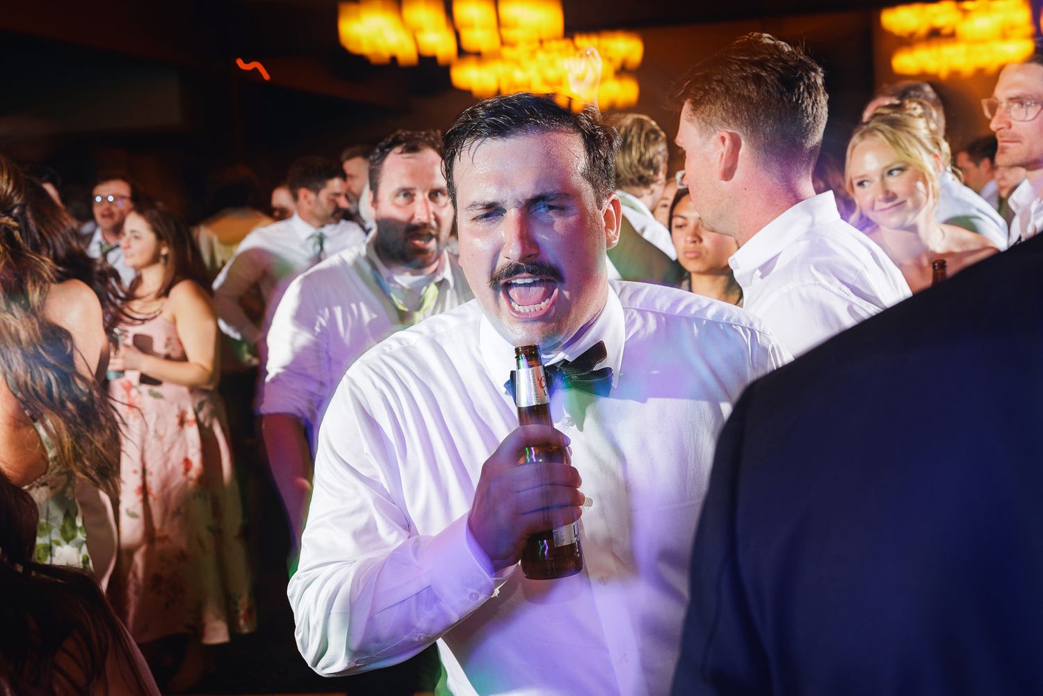 Excited male wedding guest with mustache holds beer bottle on crowded dance floor.