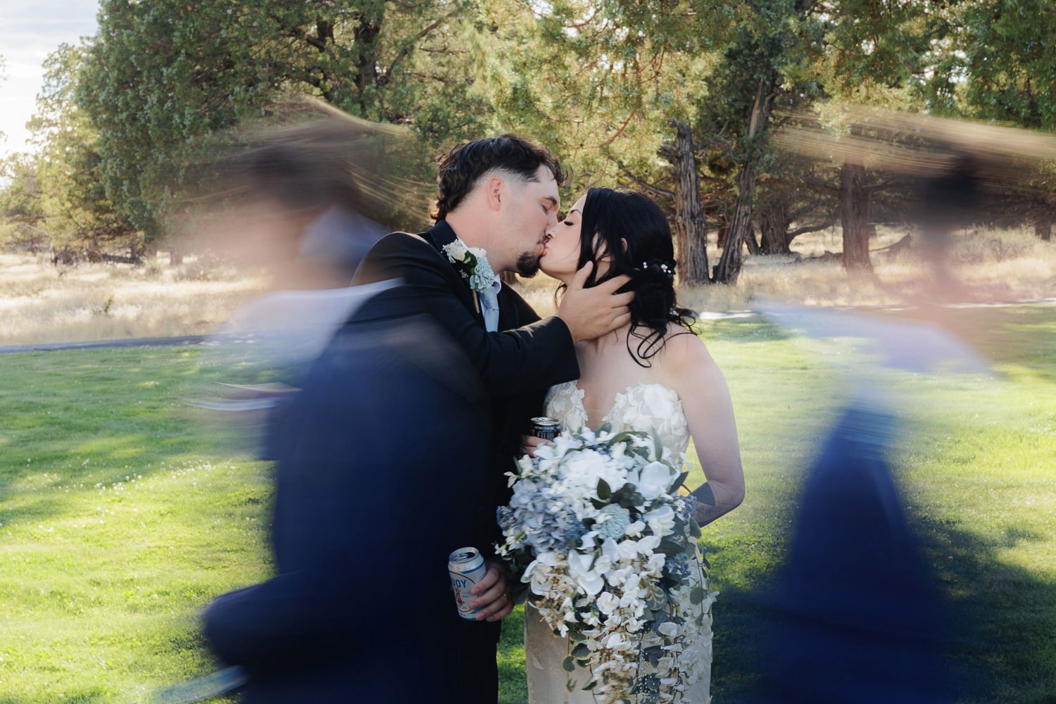 A romantic wedding couple shares a kiss on a sunny day in a park with a beautiful blue floral bouquet.