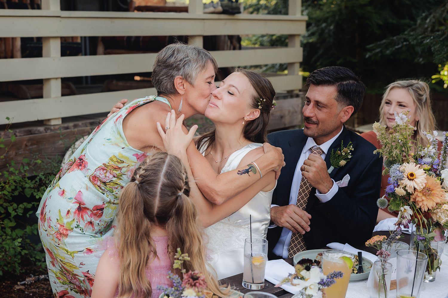 A joyful moment at an outdoor wedding reception as guests share hugs and laughter around a decorated table.
