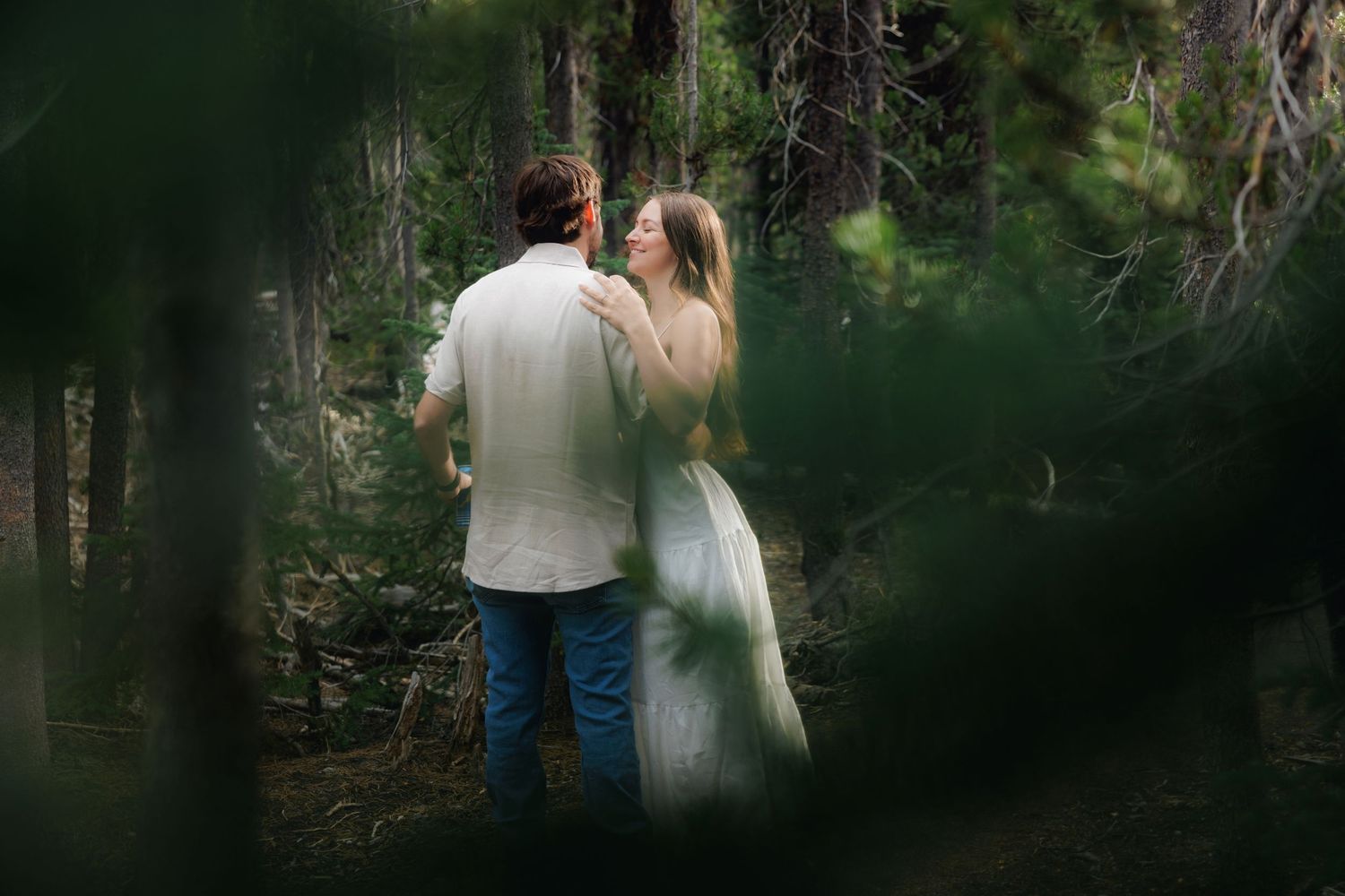 A couple in romantic embrace photographed through foliage in a dreamy forest setting.