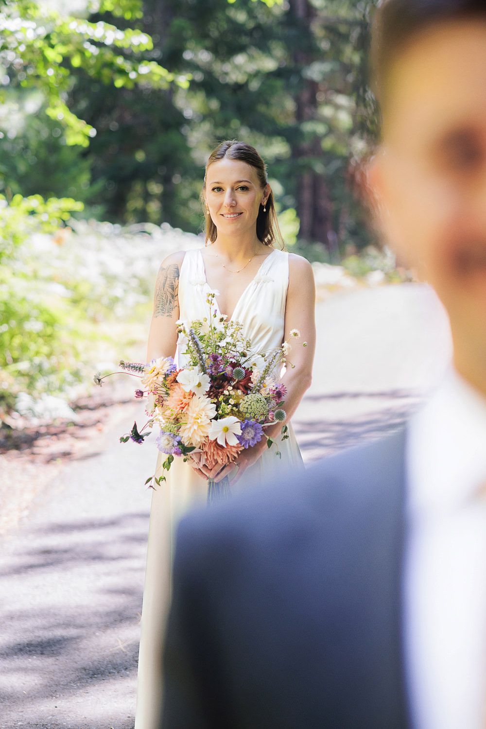 Someone in a white v-neck wedding dress holds a colorful wildflower bouquet while standing on a sunlit forest path.