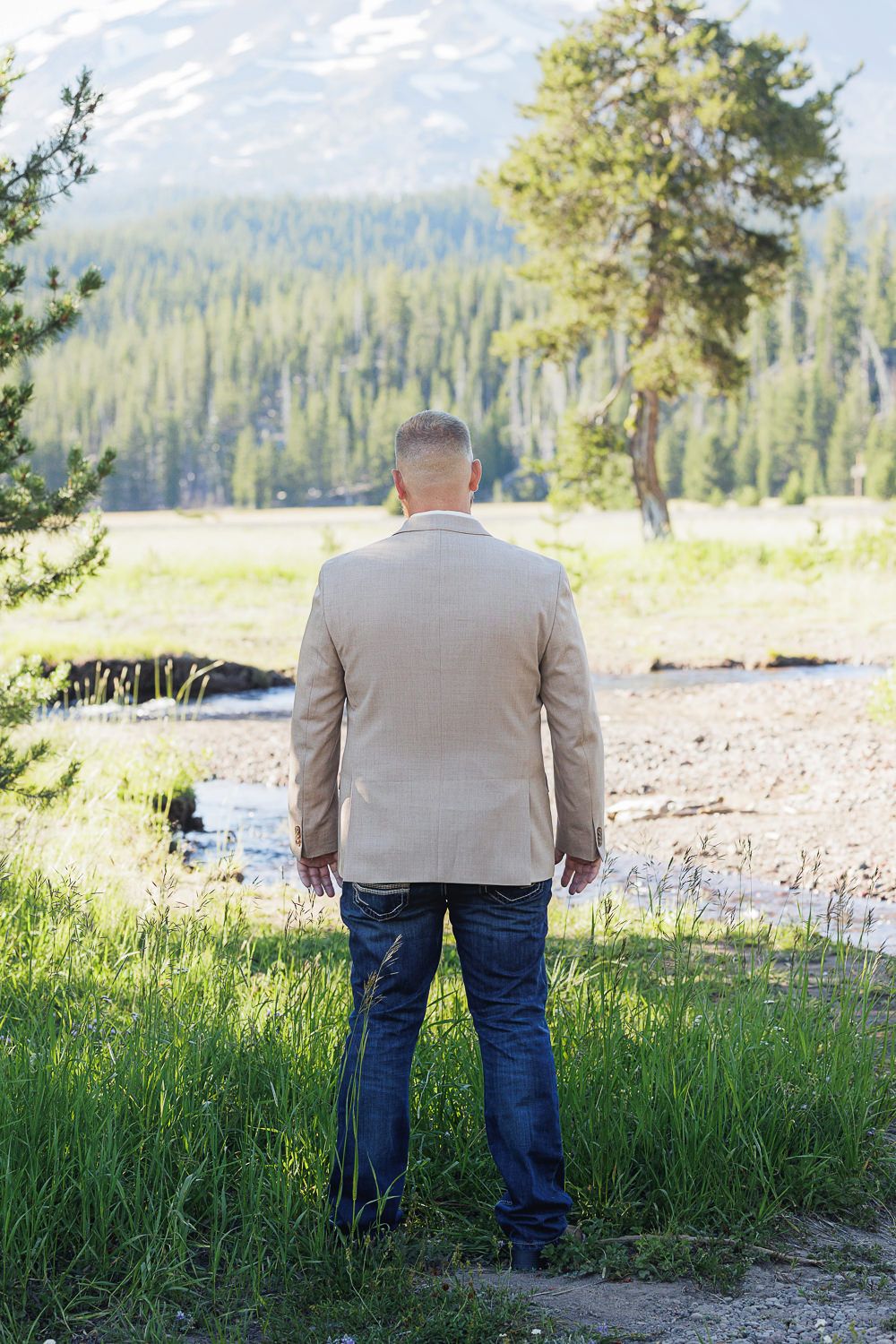 Someone wearing beige jacket and jeans stands overlooking a scenic mountain forest landscape with a stream nearby.