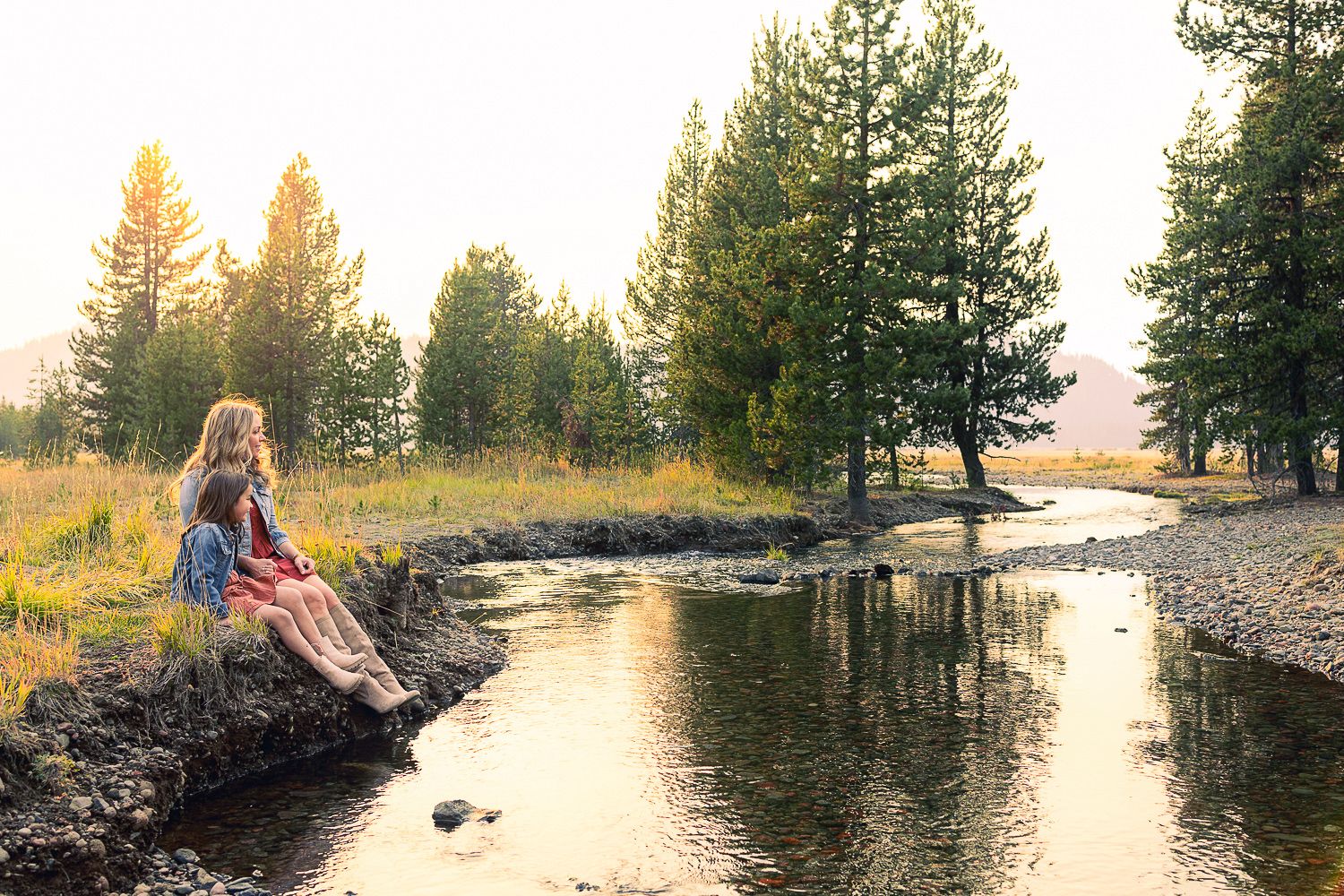 A person sits on a rocky shoreline at sunset as golden light filters through evergreen trees along a peaceful stream.